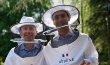 Deux hommes souriants, vêtus de combinaisons d’apiculteurs blanches avec le logo "Hédène Paris", posent côte à côte en extérieur, devant un décor verdoyant. Ils portent des chapeaux avec voiles de protection relevés, indiquant qu’ils viennent de quitter ou vont entrer dans une zone de ruches. Il s’agit des cofondateurs de la Maison Hédène, Cyril Marx et Alexis Ratouis, engagés dans la valorisation du miel français d’exception.   